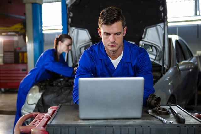 Mechanics in blue overalls working in a repair garage. One mechanic is focused on a laptop, while the other is working on a car engine. Useful for illustrating automotive repair, technology in maintenance, and teamwork in workshops.