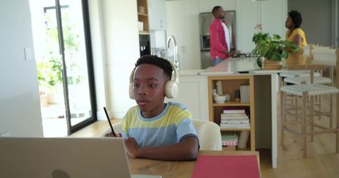 African american boy doing remote learning with headphones at home while parents talk
