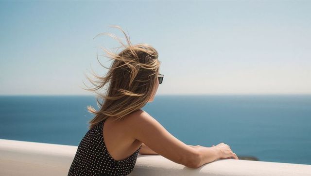 Woman gazing over calm blue ocean from white balcony with wind-blown hair and sunglasses
