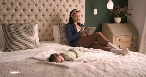 Mother Relaxing with Tablet Beside Sleeping Baby in Cozy Bedroom