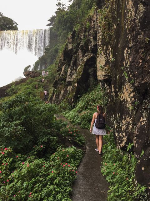 This depicts a woman with a backpack hiking along a narrow path near a breathtaking waterfall in a dense, lush forest. Appears serene and adventurous, making it perfect for travel blogs, adventure tourism promotions, outdoor gear advertisements, and nature exploration websites.