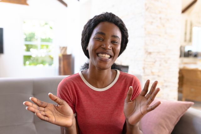Cheerful African American woman gesturing and smiling in a bright living room. Ideal for use in lifestyle blogs, home decor advertisements, and content promoting happiness and positivity. Perfect for illustrating concepts of comfort, relaxation, and joyful living.
