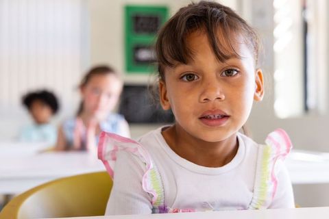 Curious elementary girl sitting at desk in bright classroom wearing pastel ruffle straps