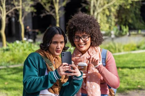 Friends comparing smartphone while laughing in park on campus with coffee and backpacks