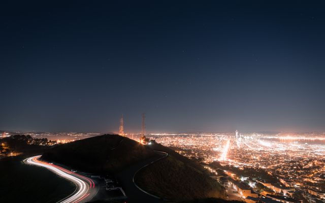 Nighttime view of a cityscape with bright twinkling lights, captured from a hilltop. Long exposure creates smooth light trails along winding roads. Silhouettes of telecommunications towers are visible against the starry sky. Perfect for use in travel websites, urban lifestyle blogs, technology themes focusing on city infrastructure, or as a background image to showcase metropolitan nightlife.