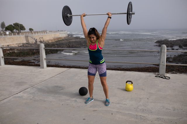 Caucasian woman lifting a barbell above her head while exercising outdoors by the seaside. She is wearing sportswear and concentrating on her strength training. A ball and kettlebell are placed behind her. This image is perfect for promoting fitness, outdoor workouts, healthy lifestyles, and strength training programs.