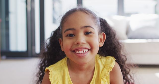 Young African American girl with curly hair smiling indoors, wearing a yellow top. Perfect for use in educational materials, family-oriented advertising, or websites focusing on children's clothing, mental health, and wellbeing. Evokes feelings of joy, playfulness, and innocence.