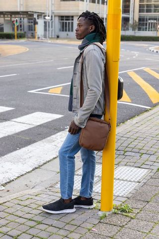 African american man standing at crosswalk leaning on yellow pole wearing denim jacket