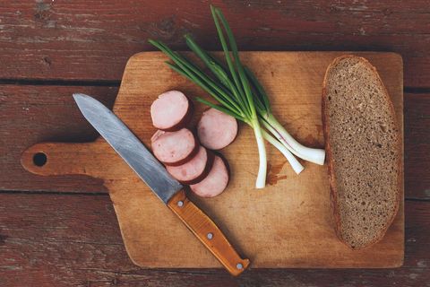 Slicing smoked sausage on rustic wooden cutting board with rye bread and green onions