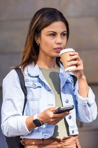 Woman sipping takeout coffee while checking smartphone in denim jacket