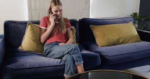 Young woman relaxing on navy velvet sofa talking on smartphone in cozy modern living room
