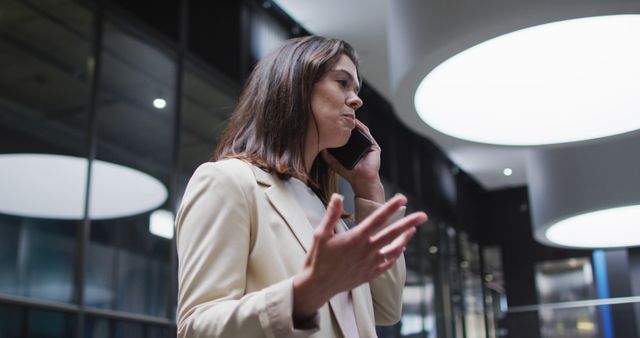 Businesswoman standing in modern office building with large windows, talking on phone and gesturing with hand. Useful for themes related to business communication, corporate work, professional environment, office concepts, and modern architecture in commercial settings.