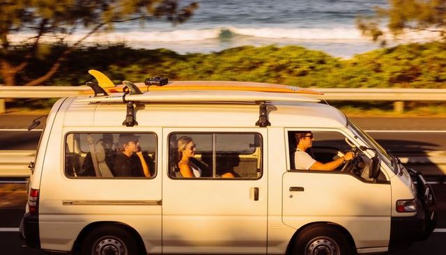 Sunlit coastal van cruising with surfboards on roof, friends wearing sunglasses
