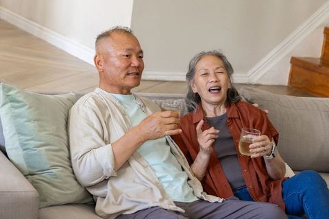 Senior asian couple relaxing on couch holding wine glasses sharing laugh at home