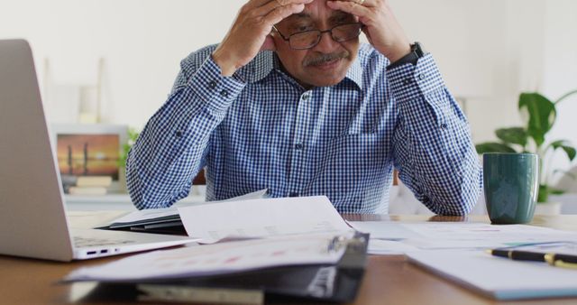 Senior man experiencing stress while managing finances at home office. Ideal for illustrating articles about financial challenges, retirement planning, personal finances, and work-life balance issues among mature adults. Can also be used in content related to home offices and remote working setups.