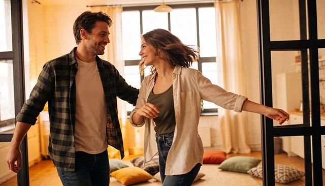 Smiling couple dancing in sunlit loft with black-framed glass door and floor cushions