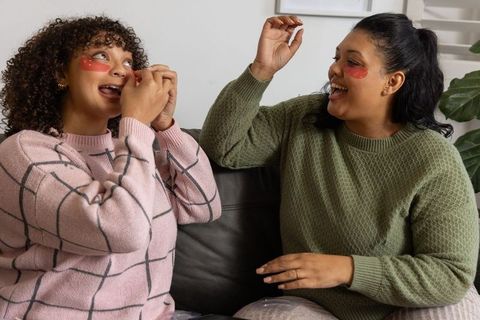 Smiling friends applying red under-eye gel patches on couch cozy home skincare routine