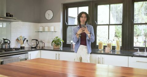 Non-binary person standing and using smartphone in sunlit modern kitchen with wood island