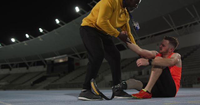 Diverse male coach helping tired disabled male athlete with running blade to stand up. professional runner training at sports stadium.