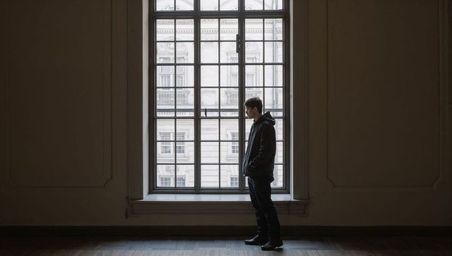 Young man standing by tall multi-pane window in empty gallery conveying urban solitude