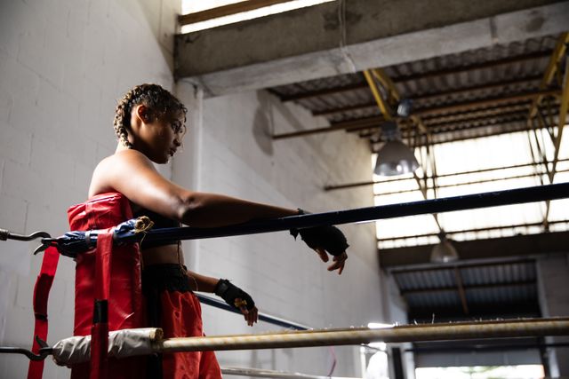 Biracial female boxer practicing in a boxing gym wearing sports clothes, resting in a corner of boxing ring. Strength sports achievement.