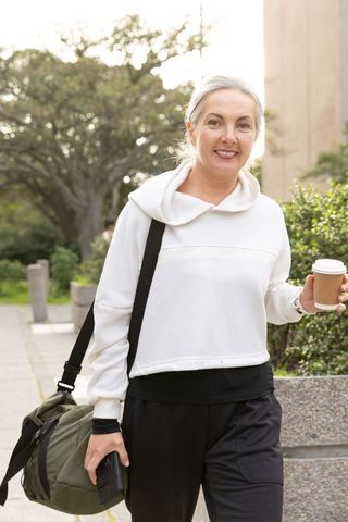Mature woman walking with coffee, duffel bag and phone in park wearing white pullover