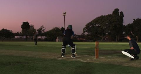 Cricket batsman batting at dusk on grassy pitch with wicketkeeper, bowler and floodlights