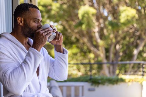 African american man sipping coffee on sunlit balcony in white bathrobe, morning calm