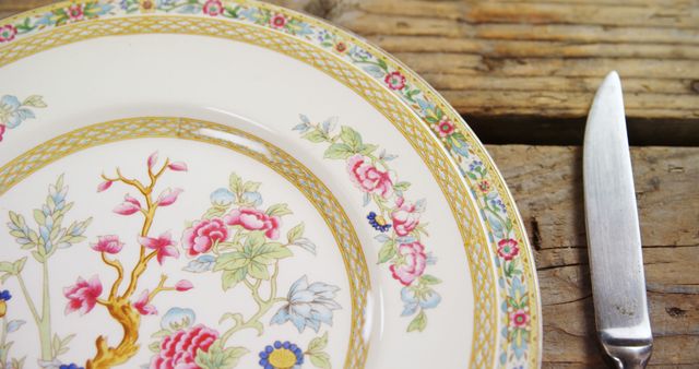 A close-up view of a decorative plate with floral patterns next to a silver knife on a rustic wooden table, with copy space. The intricate design on the plate suggests a setting prepared for a meal with a touch of elegance.