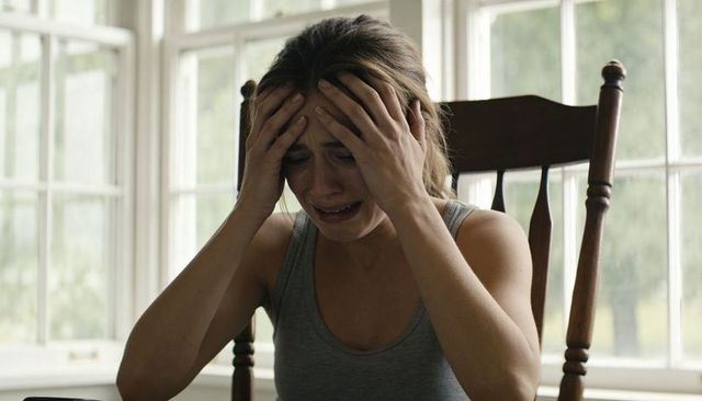 Young woman crying and clutching head on wooden chair by bright window