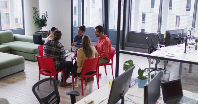 Five diverse coworkers sit in a circle discussing strategies in a modern open office. Suitable for depicting teamwork, collaboration, office culture, and contemporary work environments across business websites, blogs, and promotional materials.