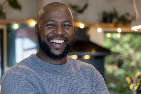 Smiling african american man wearing grey sweater in cozy kitchen with warm string lights