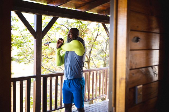 Senior African American man stretching his arms while exercising on a balcony of a log cabin. Ideal for use in content related to fitness, active lifestyle, retirement, healthy living, and outdoor activities. Can be used in articles, advertisements, or social media posts promoting wellness, solitude, and vacation retreats.