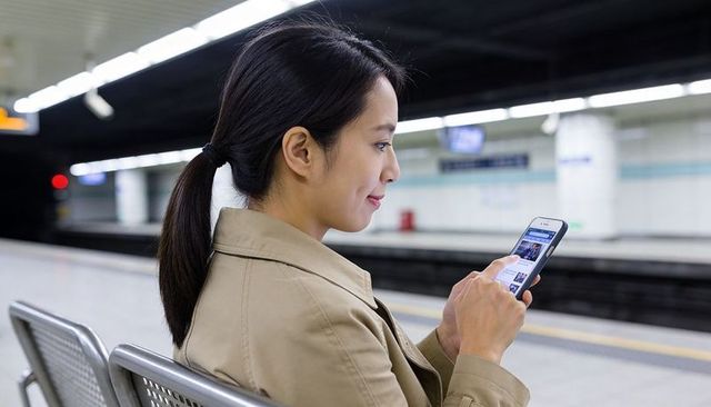 Asian commuter woman using smartphone on subway bench reading news while waiting for train