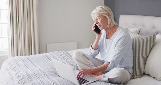 Senior woman with white hair sitting on bed, using phone and laptop. Ideal for themes on elderly life, technology use among seniors, home comfort, telecommunication, remote communication, and senior independence. Can be used in advertisements, blogs, articles, websites related to technology, senior care, home life, and communication solutions for the elderly.