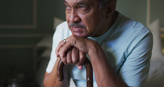 Middle-aged man leaning on a cane, gazing thoughtfully into the distance in a quiet room with dim lighting. Could be used to represent introspection, aging, solitude, or care for the elderly. Suitable for articles on mental health, senior living, aging gracefully, or elder support services.