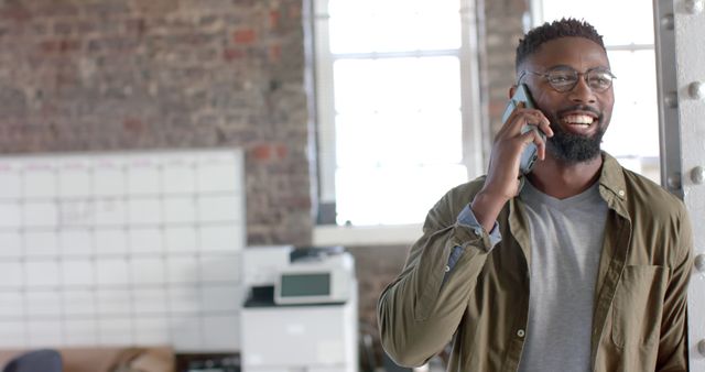 This image shows a young African American man talking on the phone in a modern office with natural light coming through large windows. The man is casually dressed and smiling, suggesting a pleasant conversation. The setting includes office furniture like a whiteboard, printer, and sofa, indicating a workspace. This stock photo can be used for business communications, advertisements for office spaces, lifestyle blogs, or technology services.