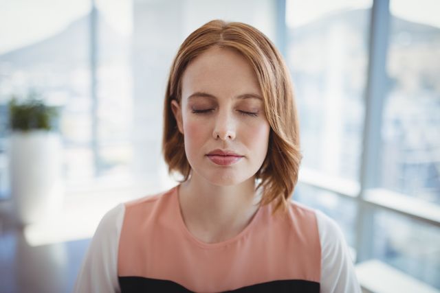 Businesswoman meditating in modern office, promoting relaxation and mindfulness in a corporate environment. Ideal for use in articles or campaigns about mental health, stress relief, work-life balance, and wellness in the workplace.