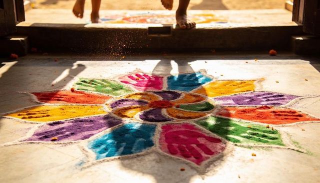 Sunlit rangoli with handprint petals at doorstep, bare feet entering festive home