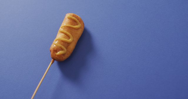 Corn dog covered in mustard on blue background. Perfect for use in ads, menu designs, food blogs, and culinary websites.