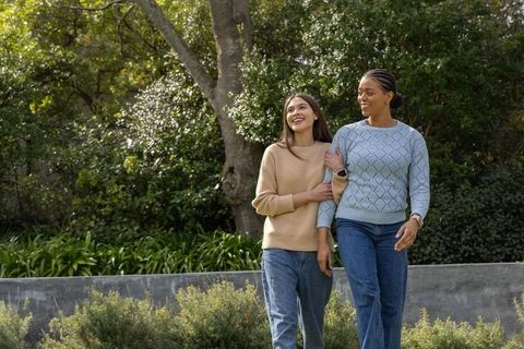 Two friends walking arm in arm through sunlit park in sweaters and jeans