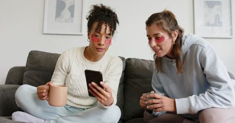 Women Sharing Cozy Self-Care Night on Couch Using Smartphone and Under-Eye Patches with Mugs
