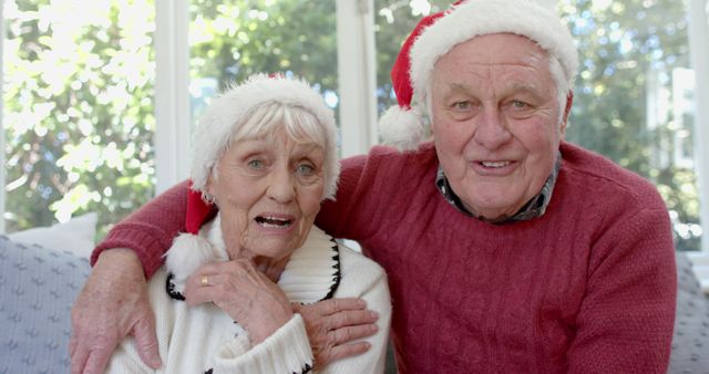 An elderly couple wearing Santa hats is celebrating Christmas indoors. The couple is hugging and looking cheerful, creating a festive and heartwarming holiday atmosphere. This stock photo is perfect for holiday greeting cards, festive invitations, and social media posts aimed at spreading holiday cheer among seniors.
