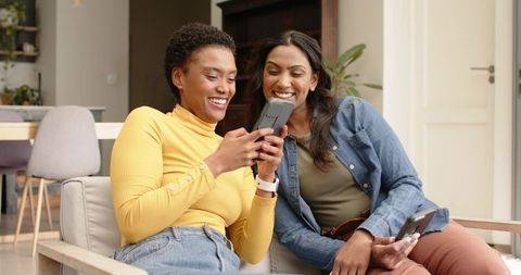 Diverse Female Friends Sharing Smartphone on Cozy Sofa Indoors