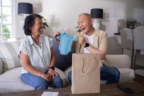 Asian senior couple celebrating baby announcement holding blue onesie and laughing