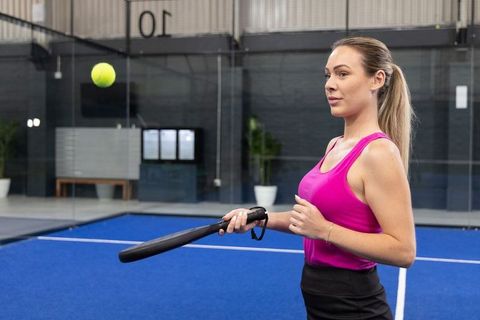 Focused woman practicing padel on modern indoor blue court holding black paddle wearing pink tank to