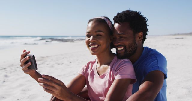 African American couple sits on sandy beach smiling and taking a selfie with smartphone. Ideal for content about love, holiday, vacations, relationships, summer activities, outdoor life, or travel promotions.