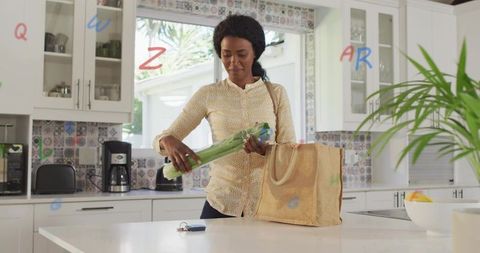 Woman packing fresh leeks into reusable tote at kitchen island, modern bright home lifestyle