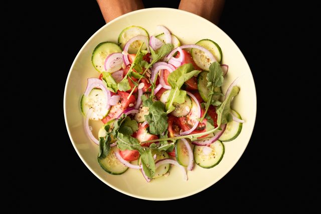 This image shows hands holding a plate of fresh vegetable salad with cucumbers, tomatoes, onions, and arugula over a black background. Ideal for use in health and wellness blogs, nutrition articles, vegan and vegetarian recipe websites, and clean eating promotions.