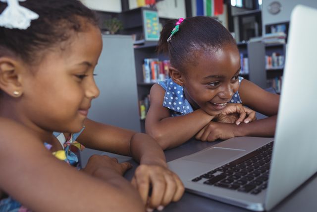 Two young girls are smiling and looking at a laptop in a library. This image can be used to depict themes of education, technology in learning, childhood friendship, and digital literacy. Ideal for educational websites, school brochures, and articles about children's learning and technology.